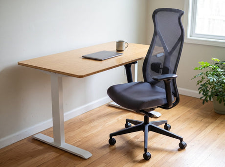 The black Nova Logix pulled up to the bamboo standing desk in a sunny home office, a closed laptop and a mug on the desk